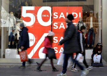 Gente che passeggia davanti ad una vetrica con il manifesto rosso con scritte bianche "Saldi 50%"