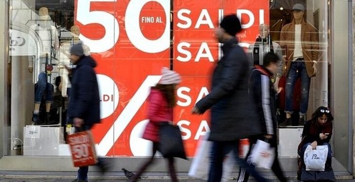 Gente che passeggia davanti ad una vetrica con il manifesto rosso con scritte bianche "Saldi 50%"