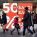 Gente che passeggia davanti ad una vetrica con il manifesto rosso con scritte bianche "Saldi 50%"